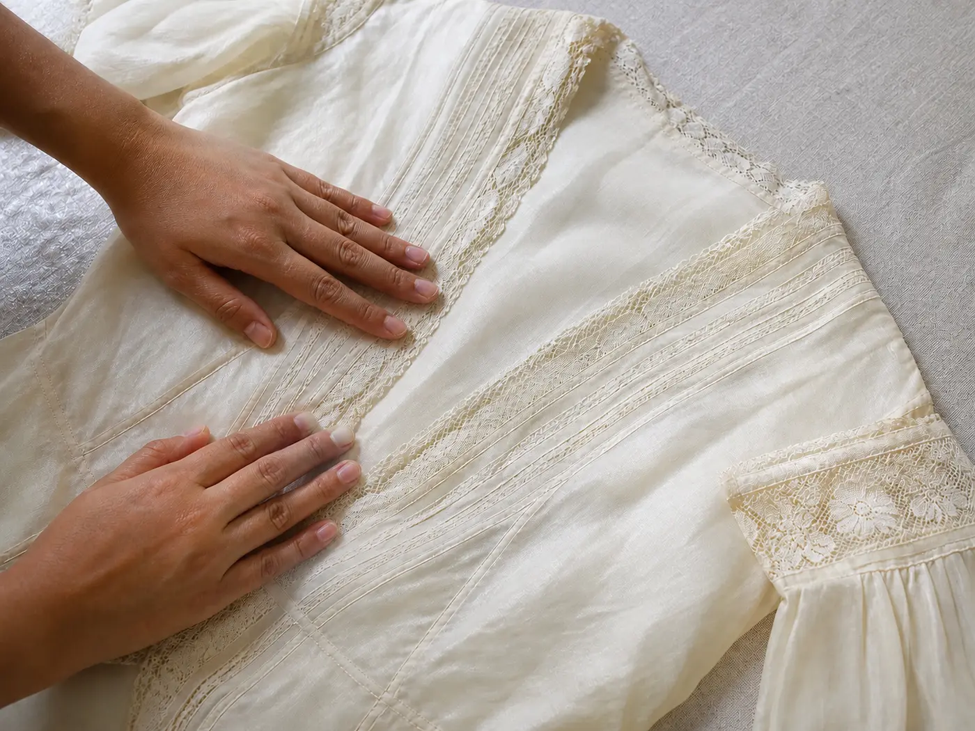 Two hands smoothing a fine vintage garment with hand-stitched detail laid flat on a pale linen bench